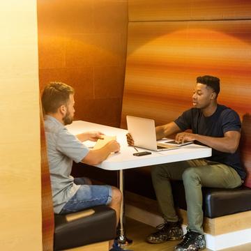 Two people in a cubby on computers talking 